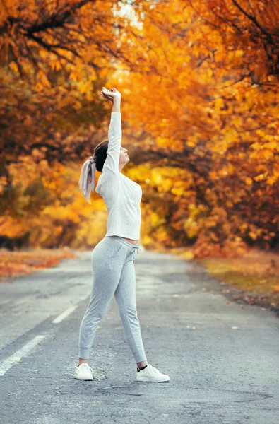 スポーツウェアの練習でスリムな若い女性とアスファルトの道路に立ち上がる手を伸ばし、女の子は秋の葉を背景に屋外スポーツに従事し、コンセプト健康的なライフスタイルと女性の美しさ — ストック写真