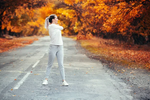 スポーツウェアの練習でスリムな若い女性とアスファルトの道路に立ち上がる手を伸ばし、女の子は秋の葉を背景に屋外スポーツに従事し、コンセプト健康的なライフスタイルと女性の美しさ — ストック写真
