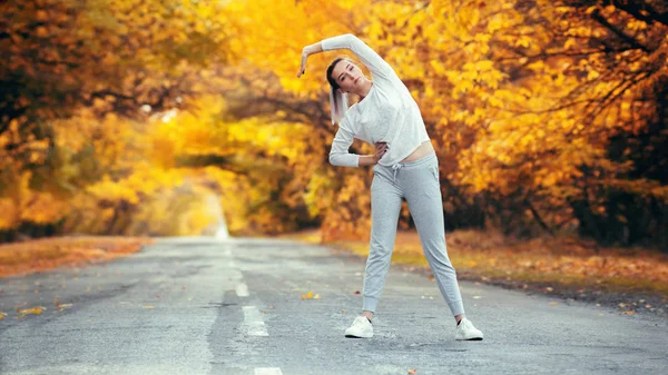 スリムな若い女性スポーツウェアの練習やアスファルトの道路での温暖化、秋の自然、コンセプトの健康とアクティブなライフスタイルを背景に屋外スポーツに従事する女の子 — ストック写真