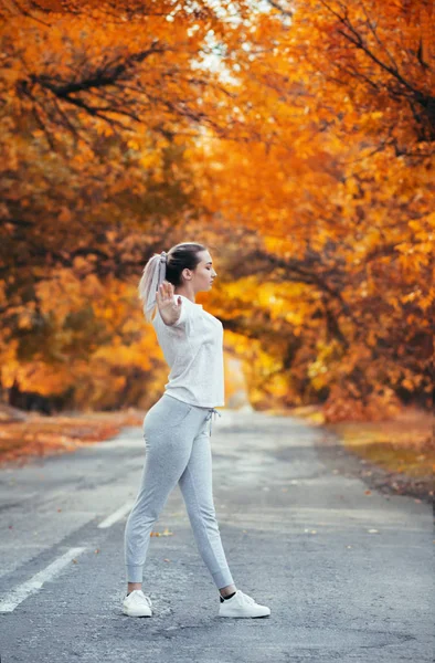 Szczupła młoda kobieta w ćwiczeniach z odzieżą sportową i rozgrzewająca się na asfaltowej drodze, dziewczyna uprawiająca sport na świeżym powietrzu na tle jesiennej przyrody, koncepcja zdrowia i aktywnego stylu życia — Zdjęcie stockowe