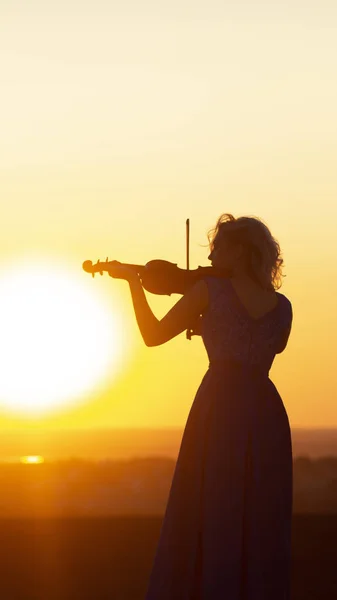 Silueta de una figura femenina en vestido largo tocando el violín al atardecer, mujer relajada en la música, actuación sobre la naturaleza, arte conceptual y hobby — Foto de Stock