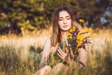 Güzel kız tarlada çiçek topluyor, uzun saçlı genç kadın doğada yürüyor, yazın köyde narin bir fotoğraf.