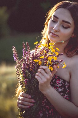 Çayırda bir buket kır çiçeğiyle güzel bir kız, uzun saçlı, doğada yürüyen genç bir kadın, köyde yazın çekilen narin bir fotoğraf.