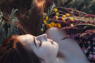 Bir buket kır çiçeğiyle çimenlerin üzerinde uzanan güzel kızın yakın plan yüzü, doğada uzun saçlı genç bir kadın, köyde yazın çekilen narin bir fotoğraf.