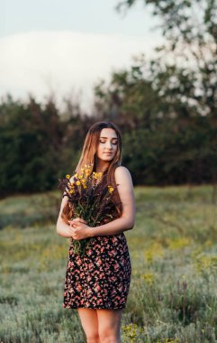 Çayırda bir buket kır çiçeğiyle güzel bir kız, uzun saçlı, doğada yürüyen genç bir kadın, köyde yazın çekilen narin bir fotoğraf.