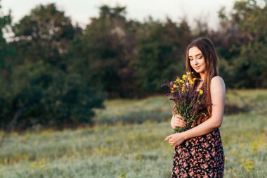 Çayırda bir buket kır çiçeğiyle güzel bir kız, uzun saçlı, doğada yürüyen genç bir kadın, köyde yazın çekilen narin bir fotoğraf.