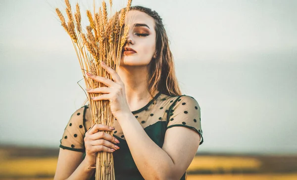 Beautiful woman face hide behind wheat ears sheaf in hand at sunset light, girl enjoying summer nature, harvest fashion