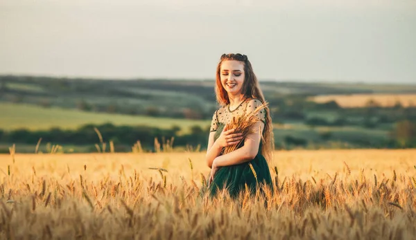 美丽的女人穿着绿色的衣服站在田里 夕阳西下手里拿着一捆麦子 女孩欣赏着夏日的自然风光 收获良多 — 图库照片