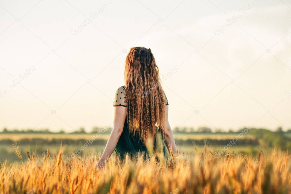 Beautiful woman in green dress walkink across field and touches ears of wheat with hand at sunset light, girl enjoying summer nature landscape