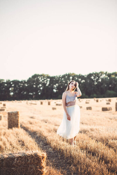 Happy young woman in dress walking in evening in field with hay bales, beautiful romantic girl with long hair outdoors in field