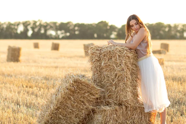 若い女性は夏の夜に干し草の上を歩くことに頼ります 日没時に野外で長い髪を持つ美しいロマンチックな女の子 — ストック写真