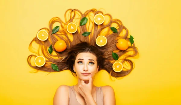 beautiful girl lying on studio background with orange fruits lay out on long hair, young woman head thinking, making choice, view from above