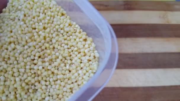 台所のボードに置かれた大きなプラスチック透明なプレート上のキビの穀物は 人間の手に触れ ベジタリアンの健康食品 テキストをコピー — ストック動画