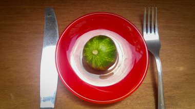 a tiny cucumber on a Red platter, but the cucumber is very similar to a small watermelon or planet Earth, next to a knife and fork, as if greedy people want to devour planet earth clipart