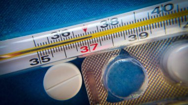 Bir cam civa termometresi, 37 derecenin üzerindeki sıcaklıkları gösteriyor, beyaz bir hap ve boş bir ilaç paketi, yukarıdan çekilmiş ve seçici odaklanmış, yakın çekim..