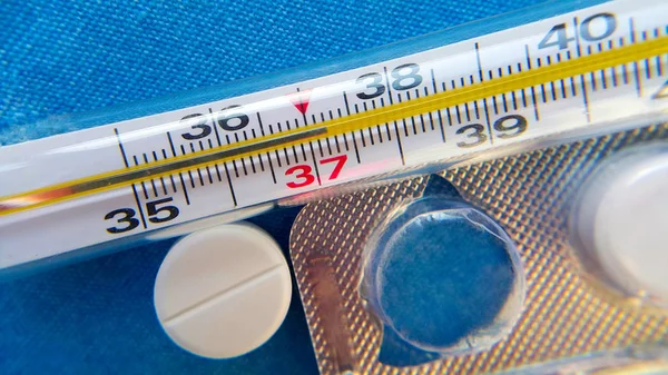 Bir cam civa termometresi, 37 derecenin üzerindeki sıcaklıkları gösteriyor, beyaz bir hap ve boş bir ilaç paketi, yukarıdan çekilmiş ve seçici odaklanmış, yakın çekim..