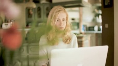 Evde bir masada oturan genç ciddi sarışın beyaz kadın izleme çekim yan yazarak yan onun laptop bakıyor