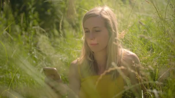 若いブロンドの白人女性は 携帯電話で自分撮りを取って 屋外の草の中に座って微笑んでいます 4Kスローモーション — ストック動画