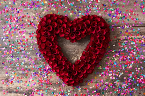 バレンタインの日に木製のテーブルの紙吹雪で飾られた赤いバラから作られた心 — ストック写真