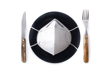 Siyah bir tabakta koruyucu yüz maskesi, çatal ve bıçak. Coronavirüs salgını sırasında restoranda yemek yemek..
