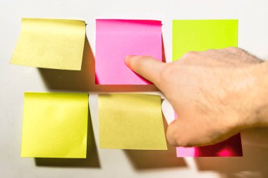 İşaret parmağı, ofisteki beyaz duvara yapıştırılmış renkli kare post-it notlarını gösteriyor.