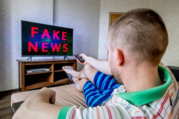 A man is sitting on a sofa and watching TV with fake news on the screen