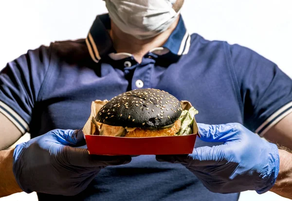 A man in a medical mask and gloves holds a black Burger in his hands on a white background