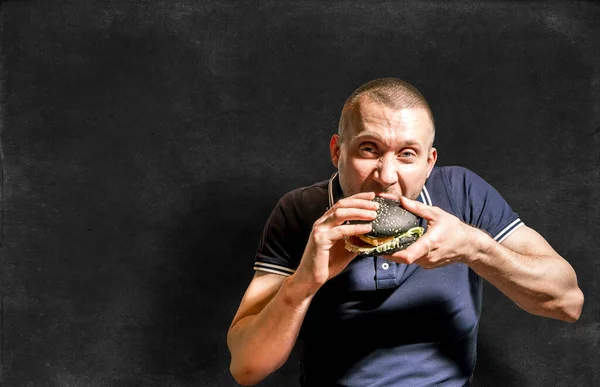 A hungry Man enjoys eating a black Burger on the background of a chalkboard.