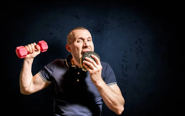 A hungry Man enjoys eating a black Burger and holding a dumbbell against a concrete wall. A man chooses between fast food and a healthy lifestyle.