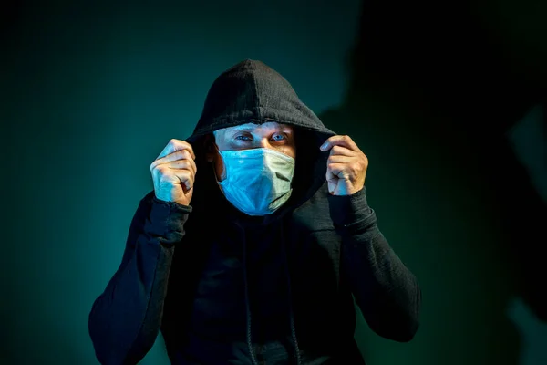 A man in a medical mask with a hood on his head poses on a dark green background