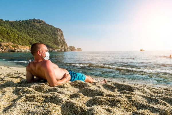 医療マスクの男が海辺のリゾートでビーチで日焼けを得る — ストック写真