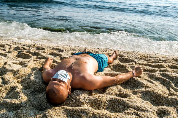 医療マスクの男が海辺のリゾートでビーチで日焼けを得る — ストック写真