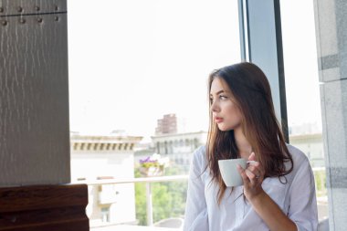 Beyaz gömlekli genç, kahverengi saçlı bir kız kapuçino kahve içiyor.