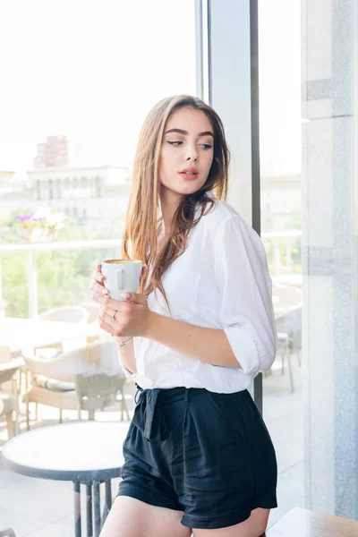카푸치노 커피를 마시는 흰색 셔츠를 입은 젊은 아름다운 갈색 머리 소녀 — 스톡 사진