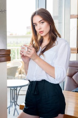 Beyaz gömlekli genç, kahverengi saçlı bir kız kapuçino kahve içiyor.