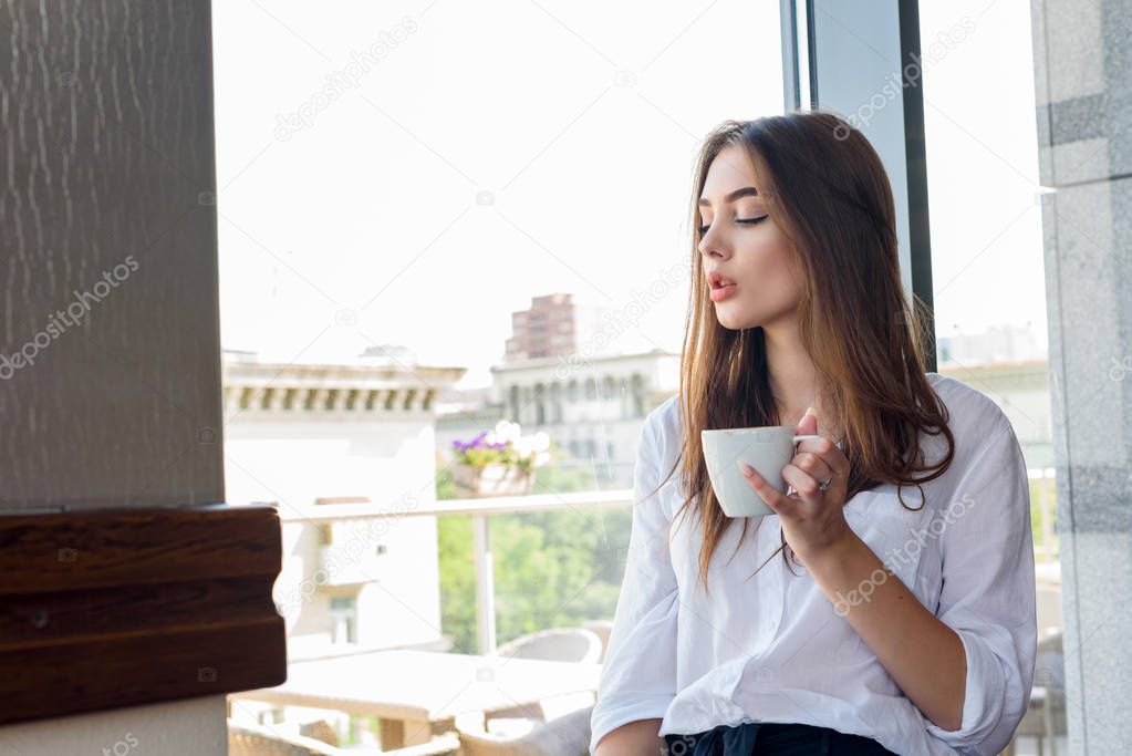 A young beautiful brown-haired girl in a white shirt drinking cappuccino coffee