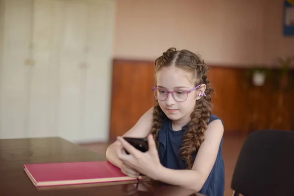 Školka sedí u stolu s učebnicích ve třídě. Dívka v modrých šatech s copánky. — Stock fotografie