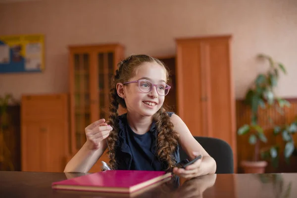 Školka sedí u stolu s učebnicích ve třídě. Dívka v modrých šatech s copánky. — Stock fotografie