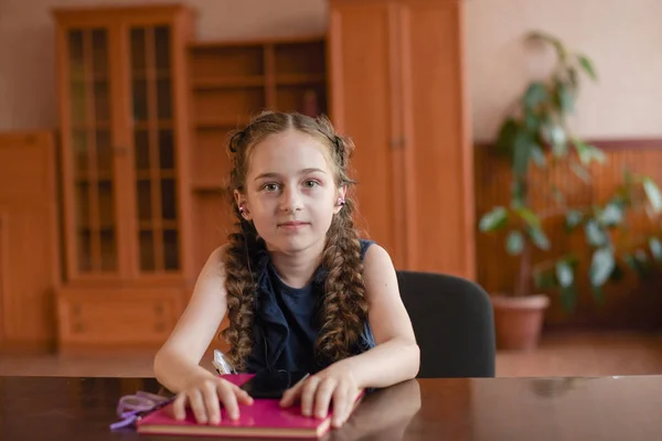 Skolflicka sitter vid bordet med läroböcker i klassrummet. Flicka i en blå klänning med Pigtails. — Stockfoto