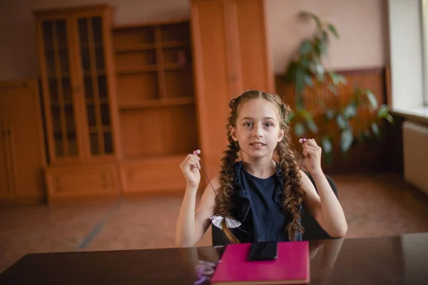 Colegiala sentada en la mesa con libros de texto en el aula. Chica en un vestido azul con coletas . —  Fotos de Stock