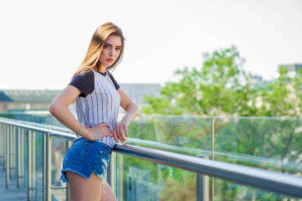 Ragazza in pantaloncini sul balcone in un bar. Ritratto estivo di giovane ragazza elegante in posa sul balcone, mini pantaloncini di jeans — Foto Stock