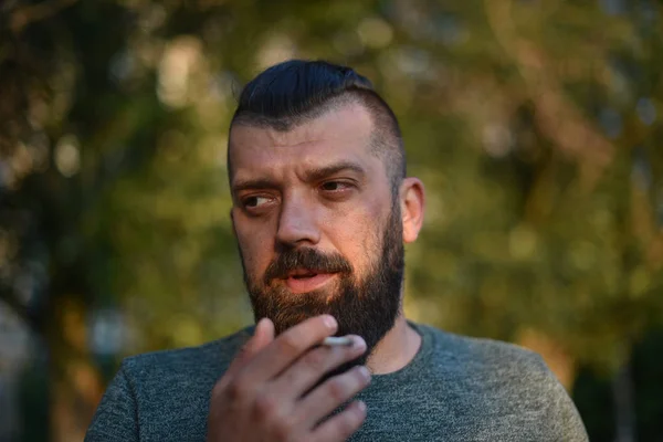 Ein Mann mit Bart raucht in einem grünen T-Shirt in der Natur eine Zigarette — Stockfoto