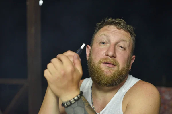 A man with a red beard smokes an electronic cigarette.