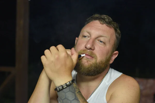A man with a red beard smokes an electronic cigarette.