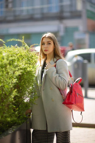 穿着灰色外套的时尚年轻漂亮女孩看着镜头，在城市的背景上微微地微笑. — 图库照片