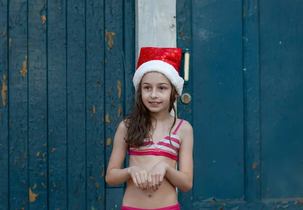 Una niña se está divirtiendo y jugando en la víspera de Navidad en el sombrero de Santa Claus, en un traje de baño, un fondo de azul . —  Fotos de Stock