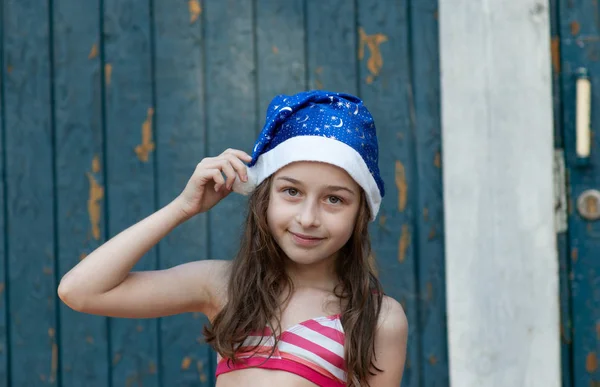 O fetiță se distrează și se joacă în ajunul Crăciunului la pălăria lui Moș Crăciun, într-un costum de baie, un fundal de albastru . — Fotografie, imagine de stoc