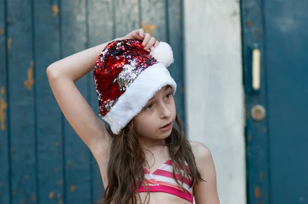Una niña se está divirtiendo y jugando en la víspera de Navidad en el sombrero de Santa Claus, en un traje de baño, un fondo de azul . — Foto de Stock