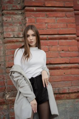 Gri paltolu genç kız bir şehir sokağının arka planında duruyor. Gri arka planda gri ceketli kız..