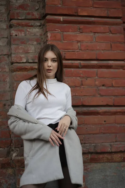 Niña con un abrigo gris se encuentra en el fondo de una calle de la ciudad. Chica en un abrigo gris sobre un fondo gris . —  Fotos de Stock